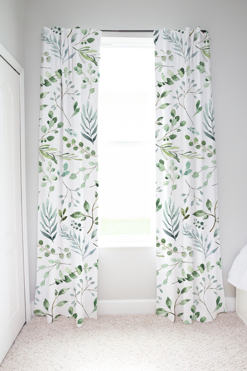 May include: White window curtains with a watercolor botanical pattern of green leaves and branches. The curtains are hanging in front of a window, and the design is repeated across both panels. The curtains add a touch of nature to the room.