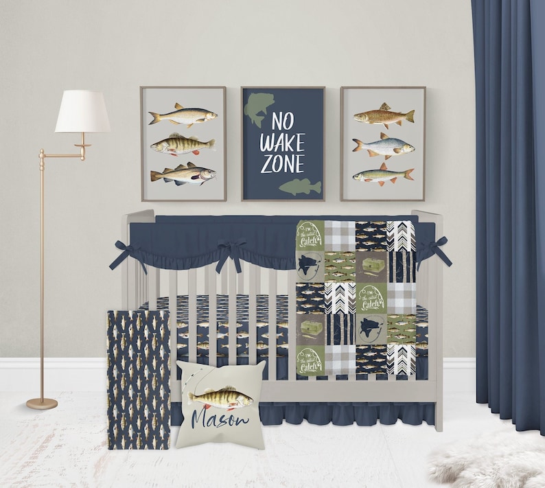 May include: A baby crib bedding set with a blue and green patchwork quilt featuring fish and the text "Catchin' Z's". The crib also has a blue and white striped bumper, a blue and white striped fitted sheet, a blue and white striped crib skirt, and a blue and white striped throw blanket. The crib is decorated with a personalised pillow with the name "Mason" and a fishing rod graphic. The crib is also decorated with three framed prints of fish and one framed print with the text "No Wake Zone".