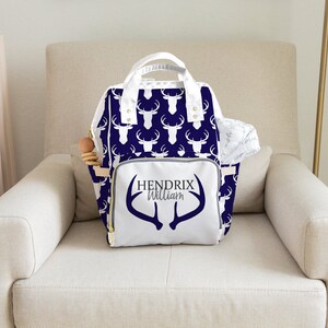 May include: A white diaper bag with a navy blue deer print and the name "Hendrix William" on the front. The bag has a white zipper and gold accents.