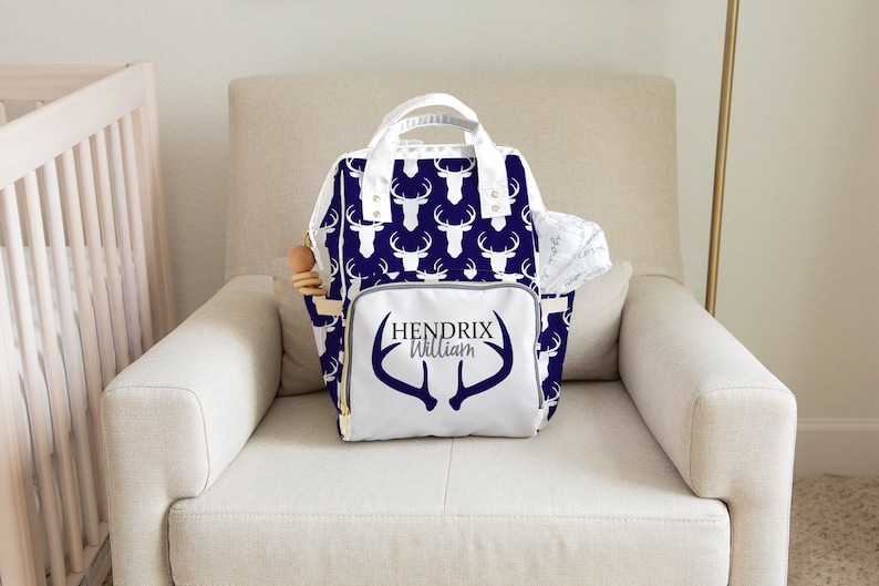 May include: A white diaper bag with a navy blue deer print and the name "Hendrix William" on the front. The bag has a white zipper and gold accents.