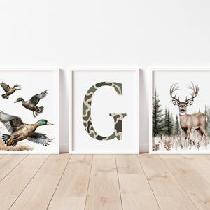 May include: Three framed art prints. One features ducks in flight, another a camouflage letter "G", and the last a deer in a forest. The prints have white frames and are displayed on a light wood floor.
