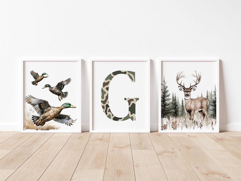 May include: Three framed art prints. One features ducks in flight, another a camouflage letter "G", and the last a deer in a forest. The prints have white frames and are displayed on a light wood floor.
