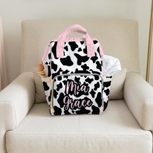 May include: A black and white cow print nappy bag with pink accents and the name "Mia Grace" embroidered on the front.