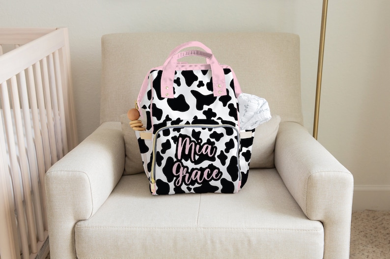 May include: A black and white cow print nappy bag with pink accents and the name "Mia Grace" embroidered on the front.