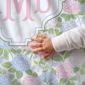 Puede incluir: Una tela blanca con un patr&oacute;n repetitivo de flores de hortensia azul claro y rosa con hojas verdes. Una letra M de monograma grande en color rosa claro est&aacute; centrada dentro de un marco decorativo con contorno azul impreso en la tela.
