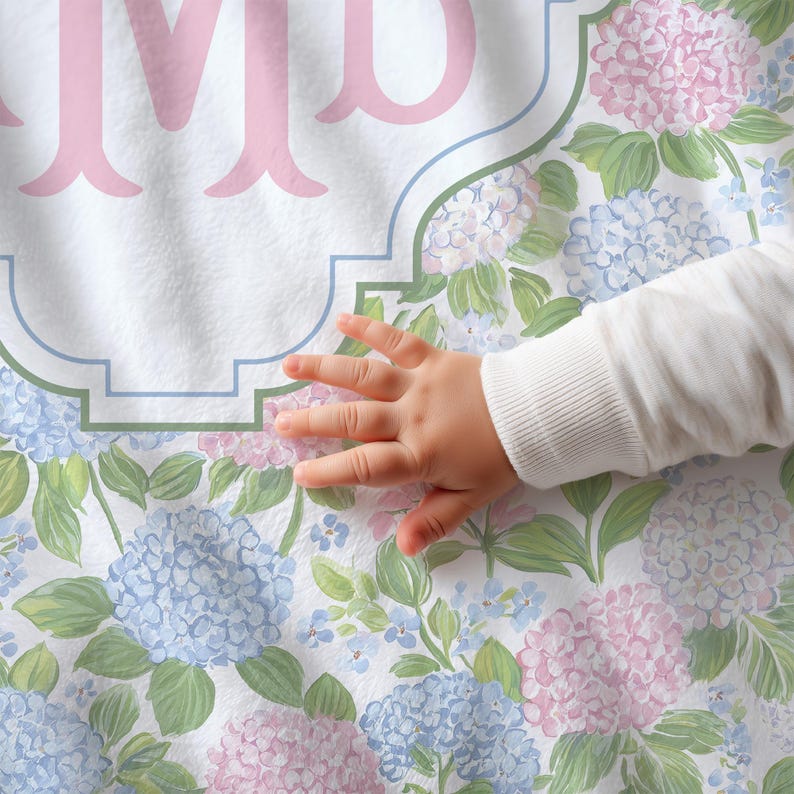 Puede incluir: Una tela blanca con un patr&oacute;n repetitivo de flores de hortensia azul claro y rosa con hojas verdes. Una letra M de monograma grande en color rosa claro est&aacute; centrada dentro de un marco decorativo con contorno azul impreso en la tela.