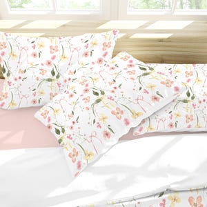 May include: Two white pillowcases with a floral pattern featuring pink and yellow flowers and green leaves. The pillowcases are on a pink and white bed.