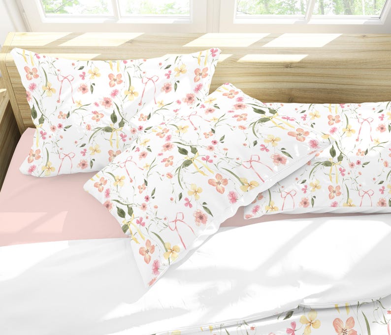 May include: Two white pillowcases with a floral pattern featuring pink and yellow flowers and green leaves. The pillowcases are on a pink and white bed.