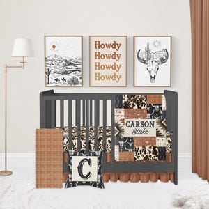 Op de afbeelding: Een zwart ledikant met een bruin en beige patchworkdeken met een zuidwestelijk ontwerp. De deken heeft de naam "Carson Blake" geborduurd. Het ledikant is versierd met een bruin en beige gepatroneerde ledikantrok en een zwart-wit kussen met een grote letter "C". Er hangen drie ingelijste prints aan de muur achter het ledikant, een met het woord "Howdy" dat vier keer wordt herhaald, een met een woestijnlandschap en een met een schedel en een cactus.