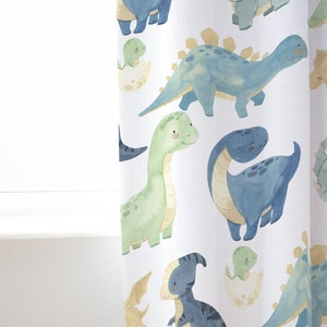 May include: White fabric with a repeating pattern of blue, green, and yellow dinosaurs. The dinosaurs are in various poses, including standing, walking, and sitting. Some dinosaurs are hatching from eggs.