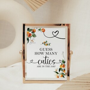 May include: A gold-framed sign with a white background and a floral design featuring orange blossoms and leaves. The sign reads "Guess how many cuties are in the jar?"