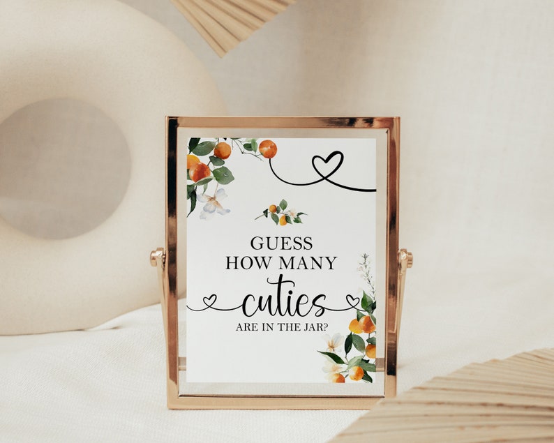 May include: A gold-framed sign with a white background and a floral design featuring orange blossoms and leaves. The sign reads "Guess how many cuties are in the jar?"