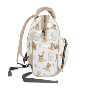 K&ouml;nnte beinhalten: Eine wei&szlig;e Wickeltasche mit einem braunen Teddyb&auml;r- und Mondmuster. Die Tasche hat einen Rei&szlig;verschluss und verstellbare Tr&auml;ger.