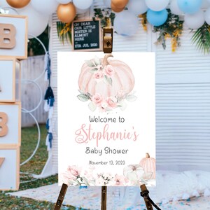 May include: A white welcome sign with a watercolor illustration of a pink pumpkin with floral accents. The text reads "Welcome to Stephanie's Baby Shower November 12, 2022".