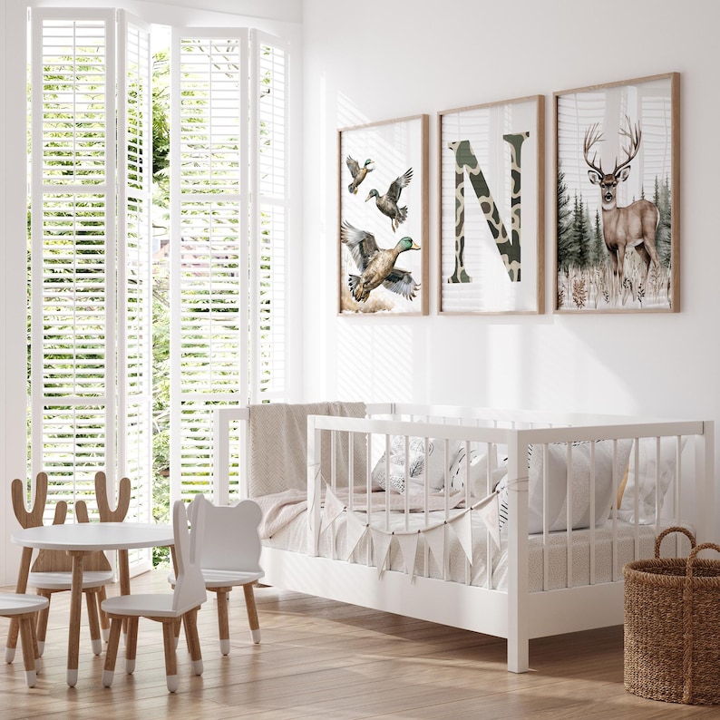 May include: A bright, airy nursery with a white crib, a small table and chairs set, and three framed prints. The prints feature ducks, the letter "N" in camouflage, and a deer. A woven basket sits on the floor.