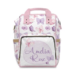 May include: A white nappy bag with a pink handle and a pink and purple watercolour butterfly pattern. The bag has a large front pocket with a pink watercolour circle and the text "Amelia Rose" in purple script.