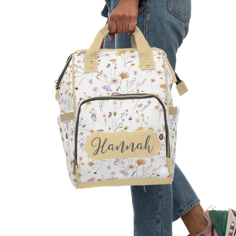 May include: A white canvas backpack with a floral print and a beige trim. The backpack has a large pocket on the front with the name "Hannah" written in a script font.