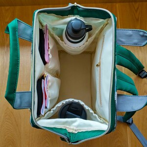 May include: A green and grey backpack with a white interior. The backpack is open and has several pockets and compartments. There are items inside the backpack, including a water bottle, a coffee cup, and some small items.
