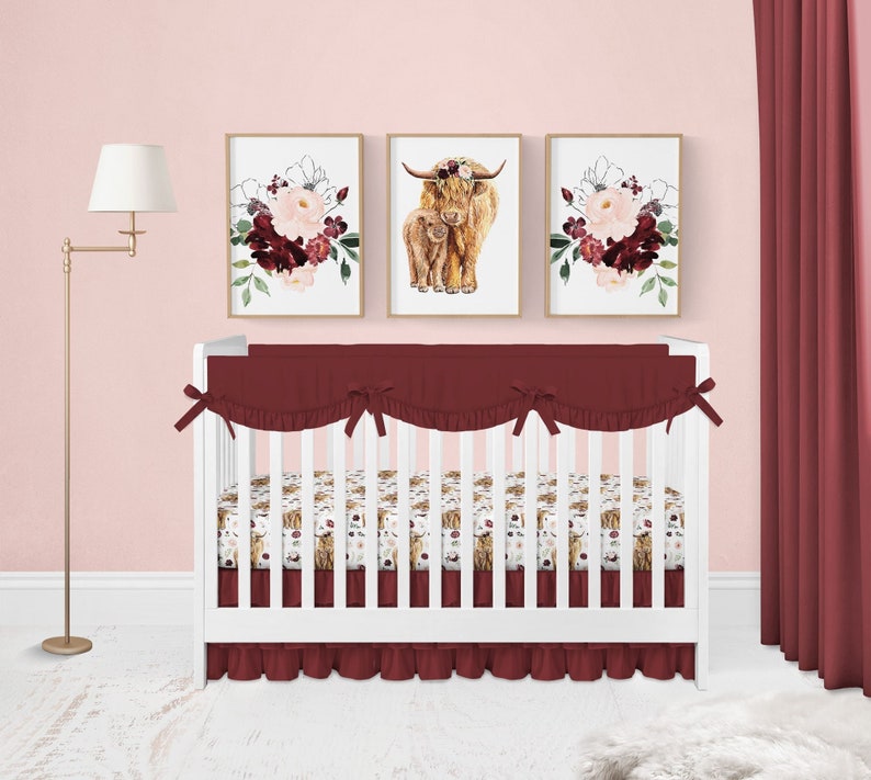 May include: A white crib with a burgundy crib skirt and a burgundy bumper. The crib is decorated with a floral and highland cow themed bedding set. Three framed prints with floral and highland cow designs are hanging on the wall behind the crib.