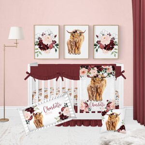 May include: A white crib with a burgundy and white floral crib bedding set. The bedding features a watercolor illustration of a Highland cow and calf with a floral crown. The crib also has a white and burgundy striped pillow with the name "Charlotte Rose" printed on it.