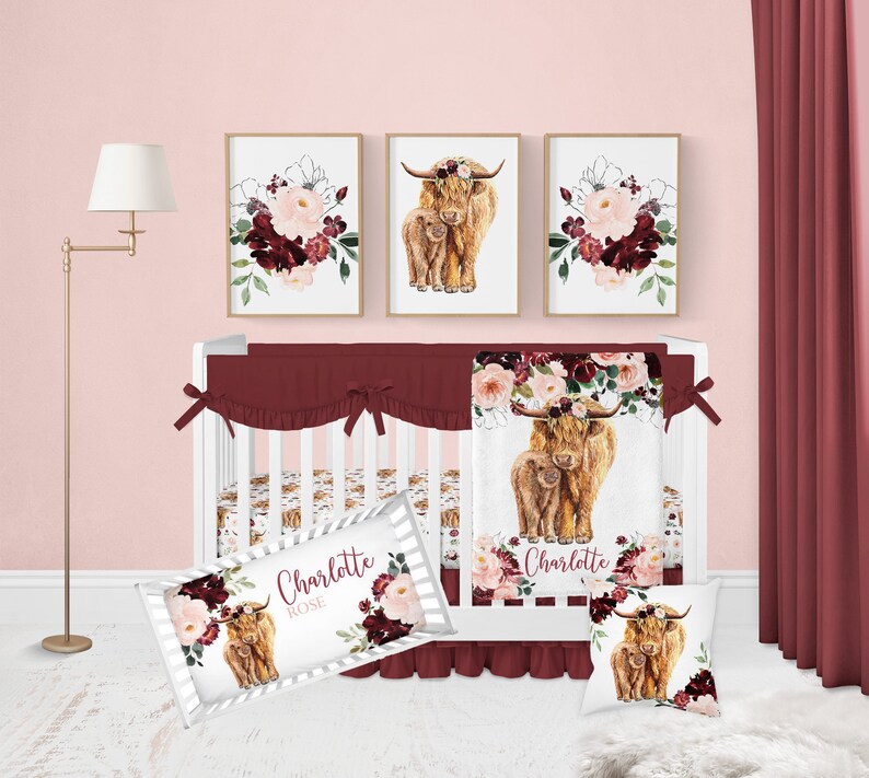 May include: A white crib with a burgundy and white floral crib bedding set. The bedding features a watercolor illustration of a Highland cow and calf with a floral crown. The crib also has a white and burgundy striped pillow with the name "Charlotte Rose" printed on it.