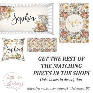 Op de afbeelding: Een bloemen wiegbeddengoedset met een witte achtergrond en een geel, oranje en roze bloemenpatroon. De set bevat een wieglaken, een kussen en een deken. Het wieglaken heeft de naam "Sophia" erop gedrukt. De deken heeft de naam "Sophia Grace" erop gedrukt. Het kussen heeft de naam "Sophia" erop gedrukt. De set is gemaakt door Little Darlings.