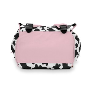 May include: A black and white cow print backpack with a pink back panel. The backpack has adjustable straps and a zipper closure.