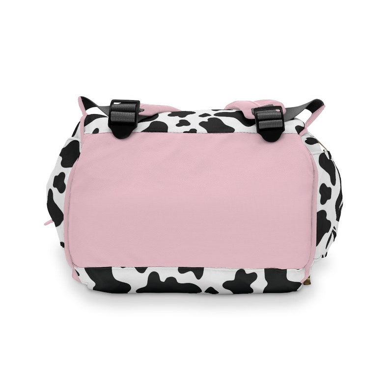 May include: A black and white cow print backpack with a pink back panel. The backpack has adjustable straps and a zipper closure.