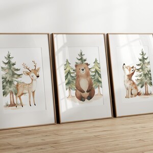 Op de afbeelding: Drie ingelijste aquarelprints met bosdieren. De eerste print toont een hert met geweien, de tweede een beer en de derde een vos. Elke print bevat groenblijvende bomen en een lichtbruine achtergrond.