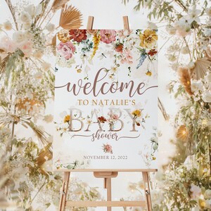 May include: A baby shower welcome sign on an easel. The sign features floral arrangements in shades of pink, yellow, and burgundy. The text reads "welcome to Natalie's baby shower" with the date November 12, 2022.