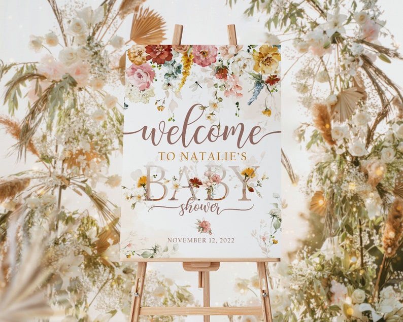 May include: A baby shower welcome sign on an easel. The sign features floral arrangements in shades of pink, yellow, and burgundy. The text reads "welcome to Natalie's baby shower" with the date November 12, 2022.