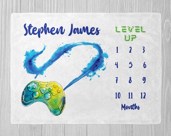 May include: A white baby milestone blanket with a blue and green watercolor design of a video game controller and the text "Stephen James LEVEL UP". The blanket has a grid of numbers 1-12 for tracking a baby's growth in months.