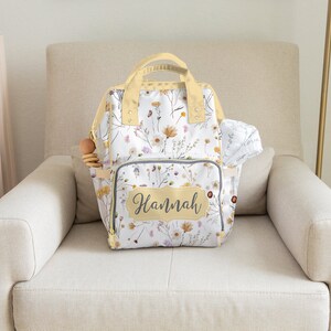 May include: A white diaper bag backpack with a floral pattern and the name "Hannah" on the front. The bag has a yellow trim and is sitting on a light beige armchair.