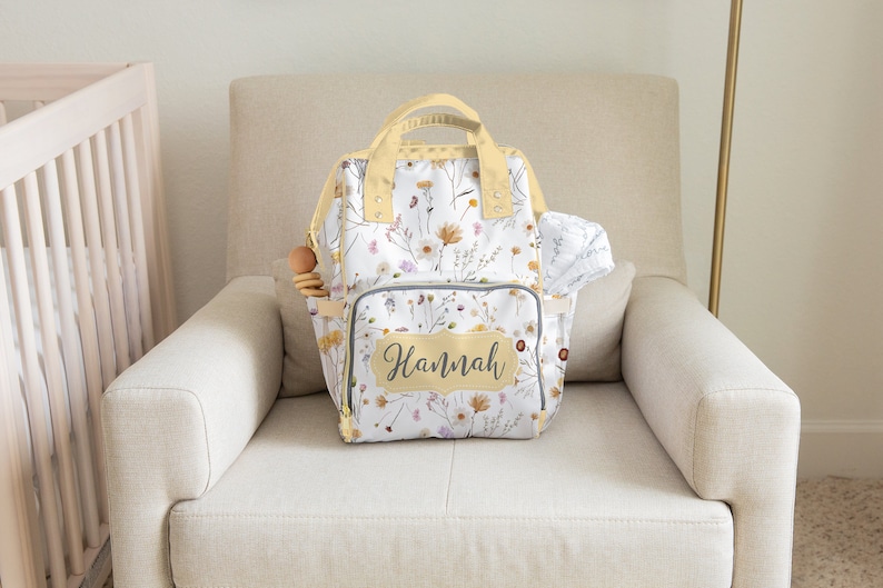 May include: A white diaper bag backpack with a floral pattern and the name "Hannah" on the front. The bag has a yellow trim and is sitting on a light beige armchair.
