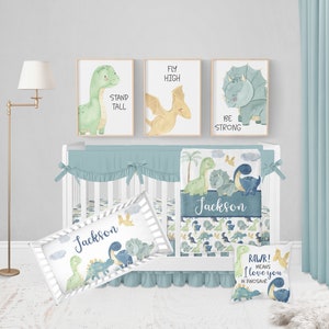 May include: A white crib with a blue and green dinosaur themed bedding set. The bedding includes a blanket, a pillow, and a bumper. The crib is decorated with three framed prints featuring dinosaurs with the text "Stand Tall", "Fly High", and "Be Strong".