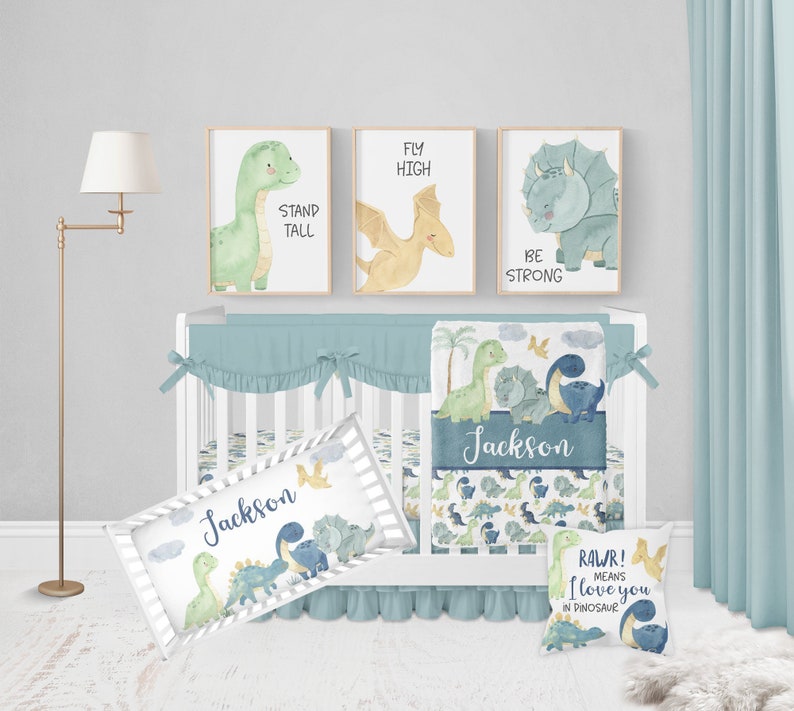 May include: A white crib with a blue and green dinosaur themed bedding set. The bedding includes a blanket, a pillow, and a bumper. The crib is decorated with three framed prints featuring dinosaurs with the text "Stand Tall", "Fly High", and "Be Strong".