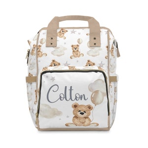 K&ouml;nnte beinhalten: Eine wei&szlig;e Wickeltasche mit einem braunen Teddyb&auml;r-Druck und dem Namen "Colton" in grauer Schrift. Die Tasche hat einen beigefarbenen Griff und Rei&szlig;verschl&uuml;sse.