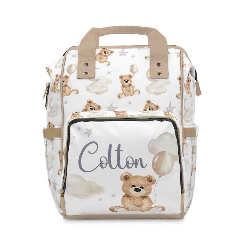 K&ouml;nnte beinhalten: Eine wei&szlig;e Wickeltasche mit einem braunen Teddyb&auml;r-Druck und dem Namen "Colton" in grauer Schrift. Die Tasche hat einen beigefarbenen Griff und Rei&szlig;verschl&uuml;sse.