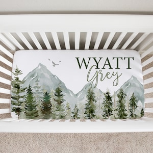 Puede incluir: Sábana de cuna blanca con una escena de montaña acuarela y árboles verdes. La sábana tiene el nombre "WYATT Grey" impreso.