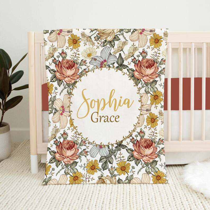 Op de afbeelding: Een wit babydekentje met een bloemenpatroon in tinten geel, oranje en groen. Het dekentje heeft een cirkel in het midden met de naam "Sophia Grace" in gouden letters.