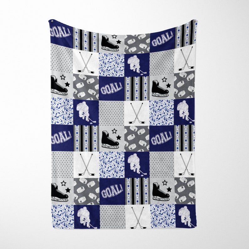 May include: A blue and gray patchwork baby blanket with a hockey theme. The blanket features hockey sticks, pucks, skates, and the word "GOAL!" repeated throughout.