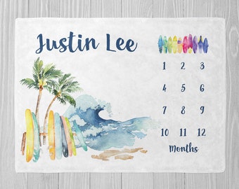 May include: A white baby milestone blanket with a watercolor illustration of palm trees, a wave, and surfboards. The blanket has a blue and white color scheme and includes the name "Justin Lee" and a numbered chart for tracking a baby's growth from 1 to 12 months.