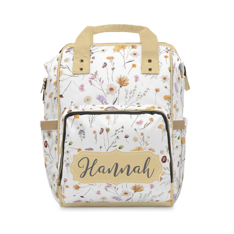 May include: A white canvas backpack with a floral print and a tan handle. The backpack has a large front pocket with a zipper and a name tag that says "Hannah".