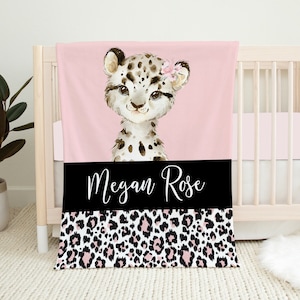 May include: Pink and black baby blanket with a watercolor illustration of a snow leopard with a pink flower. The blanket has a black stripe with the name "Megan Rose" in white cursive lettering and a pink and black leopard print pattern.