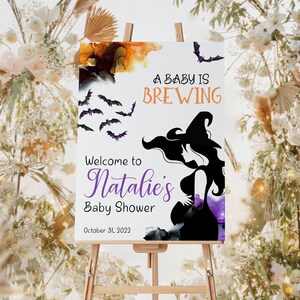 Op de afbeelding: Een wit bord met een silhouet van een heks en vleermuizen. Het bord heeft de tekst "A baby is brewing" en "Welcome to Natalie's Baby Shower" met de datum "October 31, 2023".