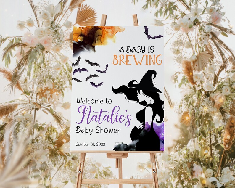 Op de afbeelding: Een wit bord met een silhouet van een heks en vleermuizen. Het bord heeft de tekst "A baby is brewing" en "Welcome to Natalie's Baby Shower" met de datum "October 31, 2023".
