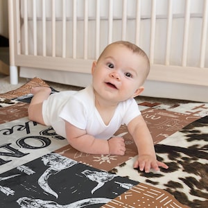 Op de afbeelding: Een baby in een wit rompertje ligt op een veelkleurige speelmat met een bruin, wit en zwart dierenprintontwerp. De mat heeft de woorden "Live, Love, Explore" erop gedrukt.