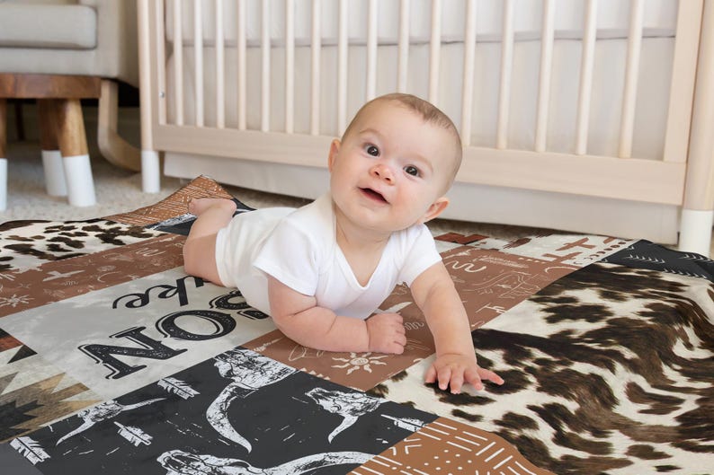 Op de afbeelding: Een baby in een wit rompertje ligt op een veelkleurige speelmat met een bruin, wit en zwart dierenprintontwerp. De mat heeft de woorden "Live, Love, Explore" erop gedrukt.