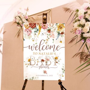 May include: A baby shower welcome sign with floral decorations in pink, yellow, and red hues. The sign is displayed on a wooden easel. The text reads "welcome to Natalie's baby shower" and the date is November 12, 2022.