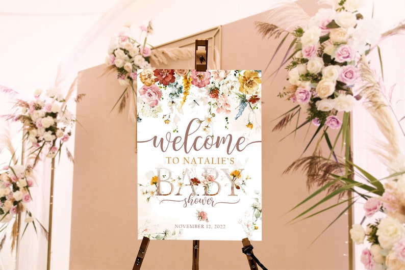 May include: A baby shower welcome sign with floral decorations in pink, yellow, and red hues. The sign is displayed on a wooden easel. The text reads "welcome to Natalie's baby shower" and the date is November 12, 2022.
